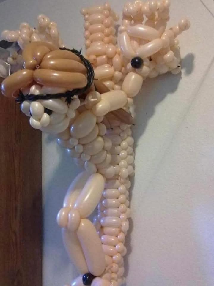 A balloon sculpture of a giraffe, crafted from light pink and tan balloons, is depicted with a black crown on its head. The…