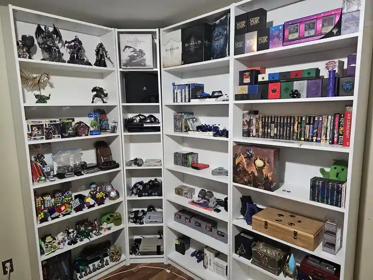 A corner of a room filled with white shelves displaying a vast collection of video games, consoles, figurines, trading card…