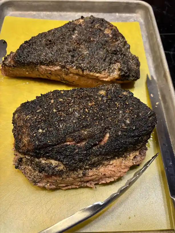 Two large, heavily charred pieces of meat sit on a yellow cutting mat inside a metal baking tray, with a knife and tongs…
