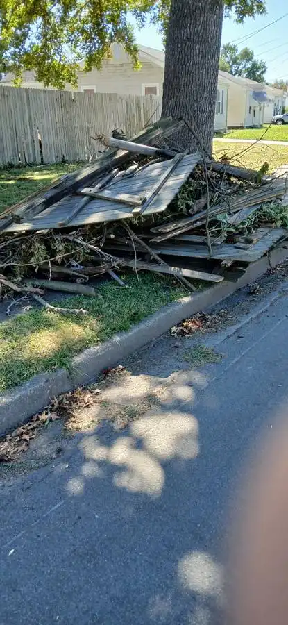 A pile of broken wooden planks and branches lies on a grassy area next to a sidewalk and tree, with a wooden fence and…