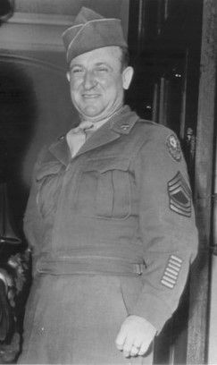 A black-and-white photo of a smiling white man in a U.S. Army uniform with sergeant stripes on his sleeve. The image has…