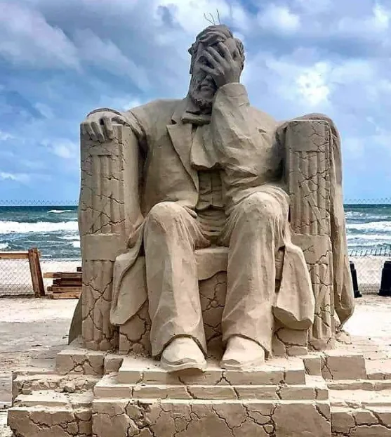 A large, detailed sand sculpture of a seated man in a suit, resembling Abraham Lincoln, is shown on a beach with ocean waves…