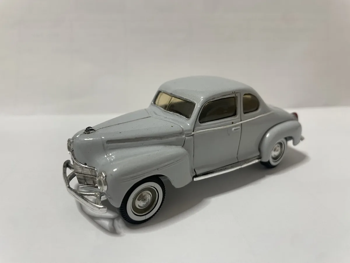 A light gray vintage-style die-cast toy car with chrome details and white-wall tires is photographed against a plain white…