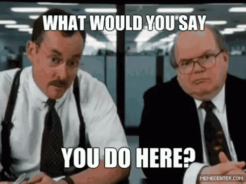 Two men in office attire sit in a cubicle environment. The man on the left, with a mustache and suspenders, looks toward the…