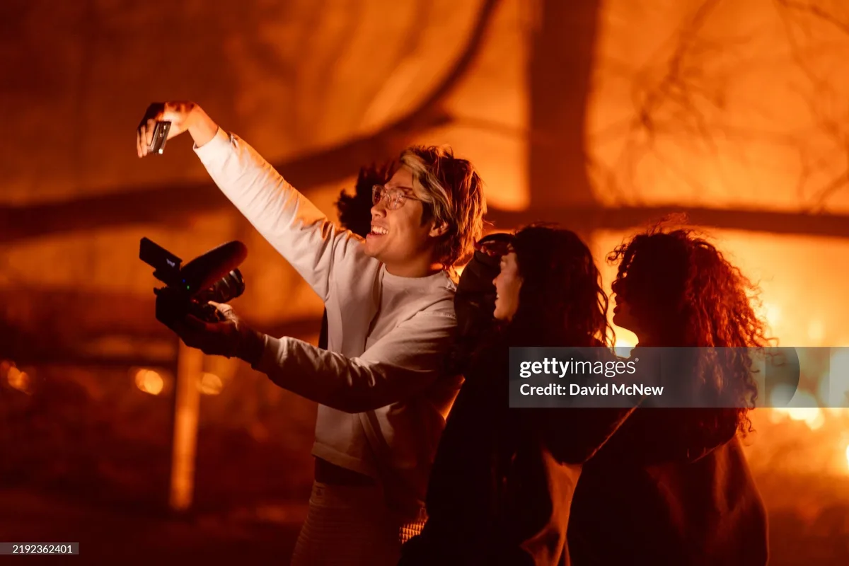 A man with glasses, wearing a white long-sleeved shirt, holds a smartphone up to take a selfie while also holding a…