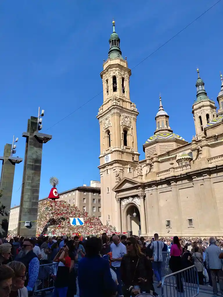 A crowded outdoor scene in front of a large, ornate church with multiple towers and a tiled dome under a clear blue sky. A…