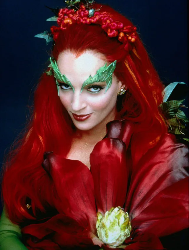 A woman with long red hair, green leafy makeup around her eyes, and a red floral costume holds a small white flower. She…