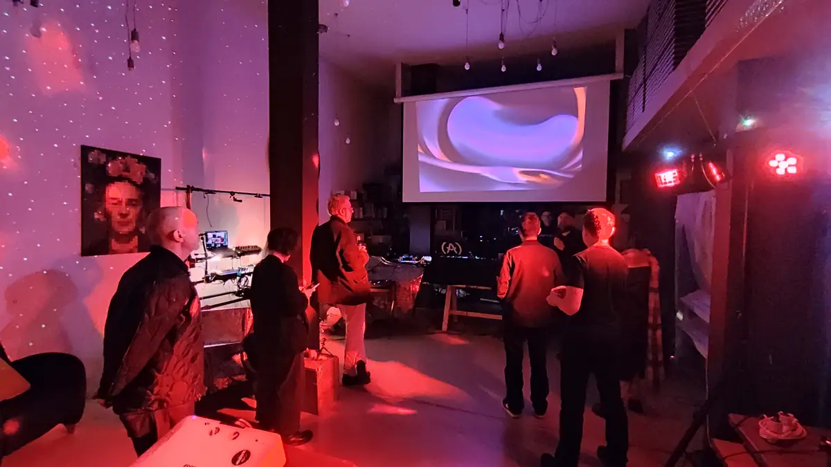 A dimly lit room bathed in red light features several people standing and watching a large projection screen displaying a…