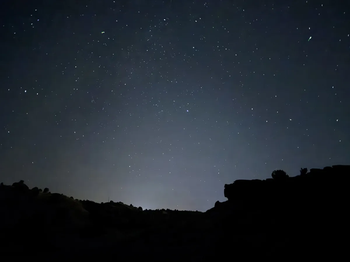 A night sky photograph showing countless stars scattered across a dark blue gradient, with the silhouette of a rocky hill or…