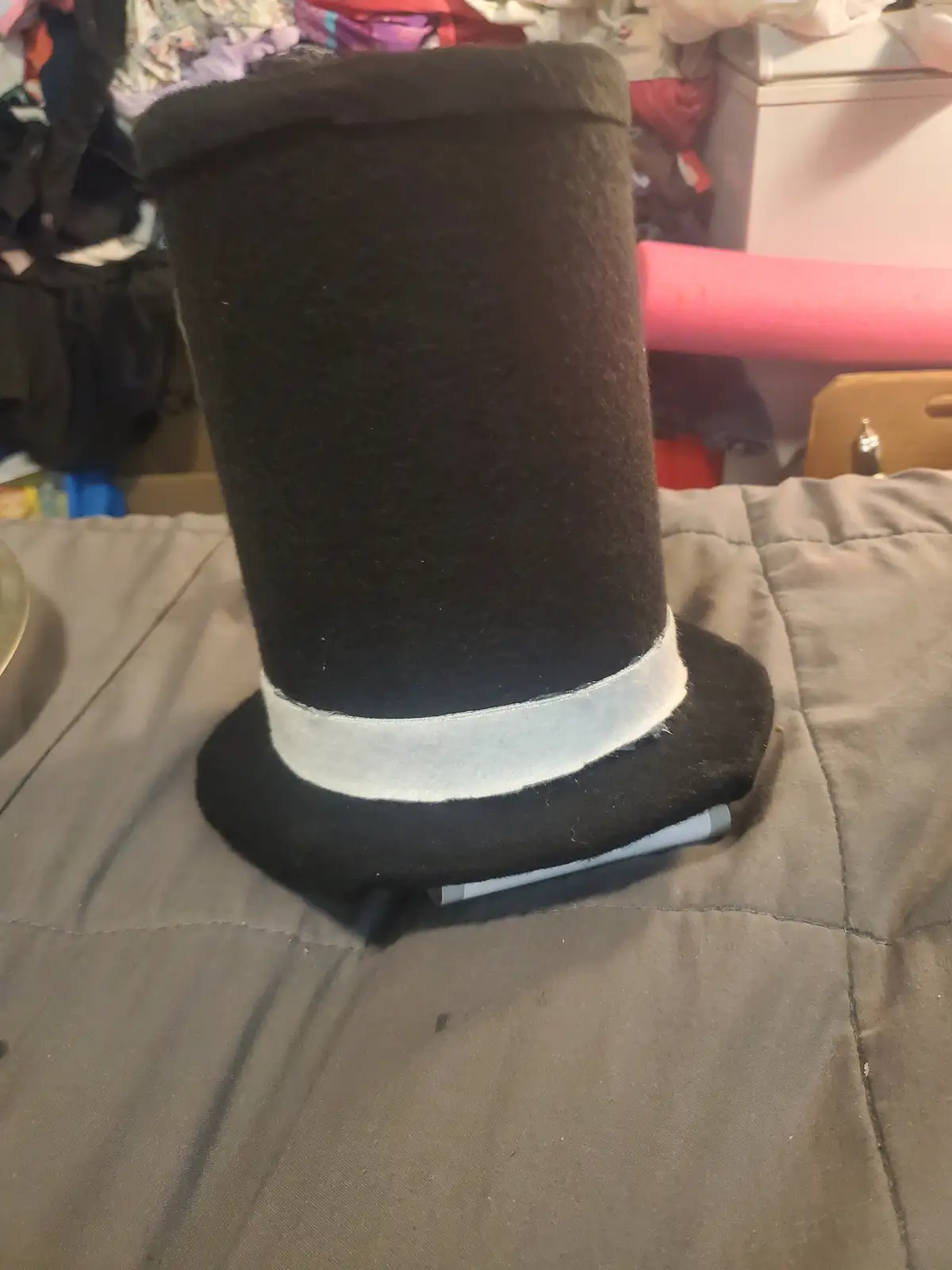 A handmade black felt top hat with a white ribbon band sits upright on a gray quilted surface, with a gray marker visibly…