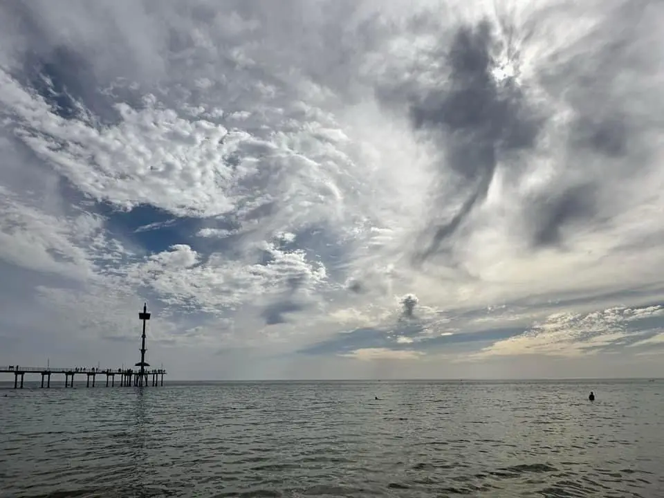 A wide-angle photo of a pier extending into the ocean under a dramatic, cloudy sky with sun rays breaking through. Several…