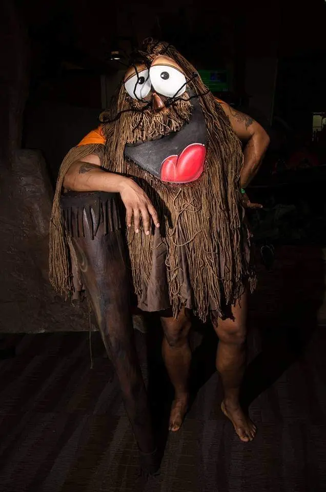 A person in a caveman costume with large cartoon eyes, a beard made of brown fibers, and a red heart-shaped mouth, poses…
