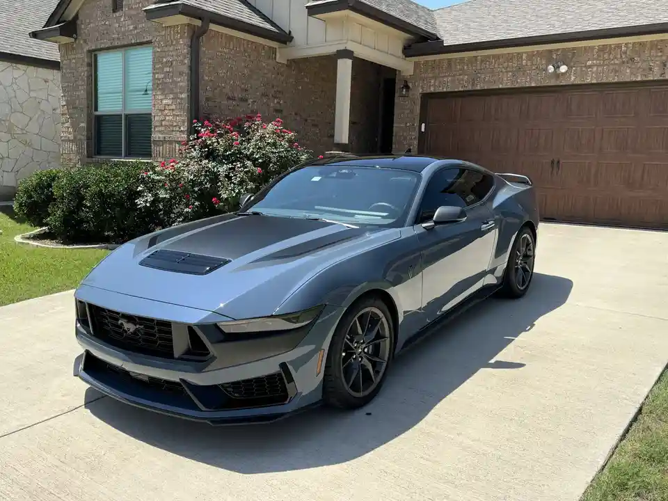 A sleek, metallic blue Ford Mustang sports car is parked on a concrete driveway in front of a modern brick house with a…
