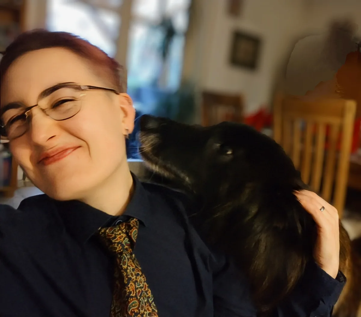 A person with short hair and glasses, wearing a blue shirt and patterned tie, smiles while holding a black dog that is…