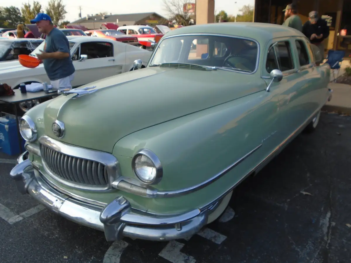 A light green vintage car with chrome details is parked at a car show, surrounded by other classic cars and people. A man in…