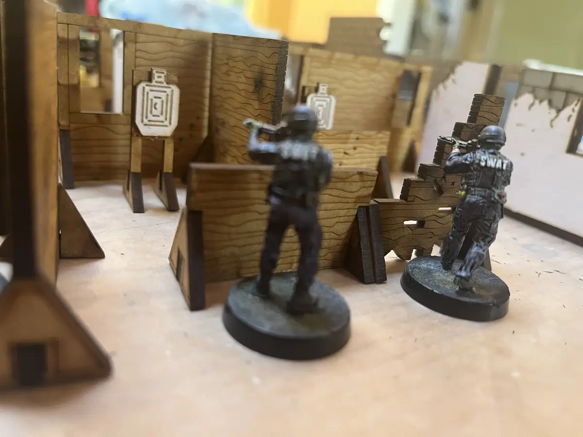Two painted miniature SWAT figures are positioned behind wooden barricades in a tabletop wargaming setup, aiming their…