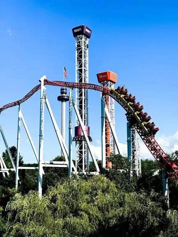 A roller coaster at Hershey's amusement park with a red track and white support structures, ascending a tall tower branded…