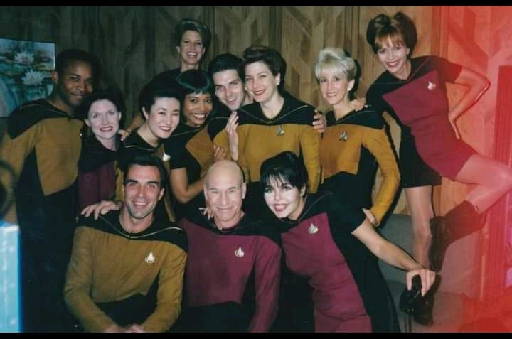a group photo of the cast of Star Trek: The Next Generation in their iconic uniforms, smiling and posing together. The crew…