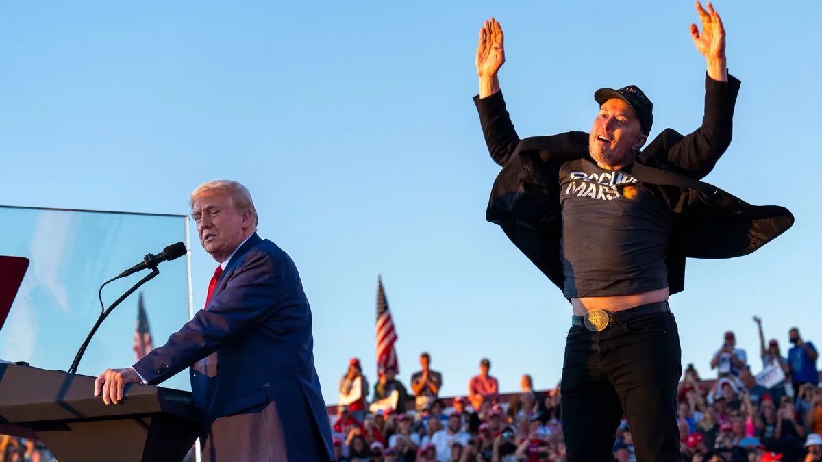A man in a suit stands at a podium speaking into a microphone while another man in a 'SpaceX' t-shirt and 'Make America…