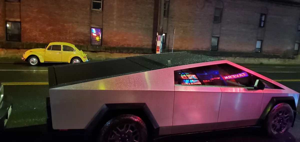 A silver, angular Tesla Cybertruck is parked on a wet city street at night, its windows displaying neon signs that read…