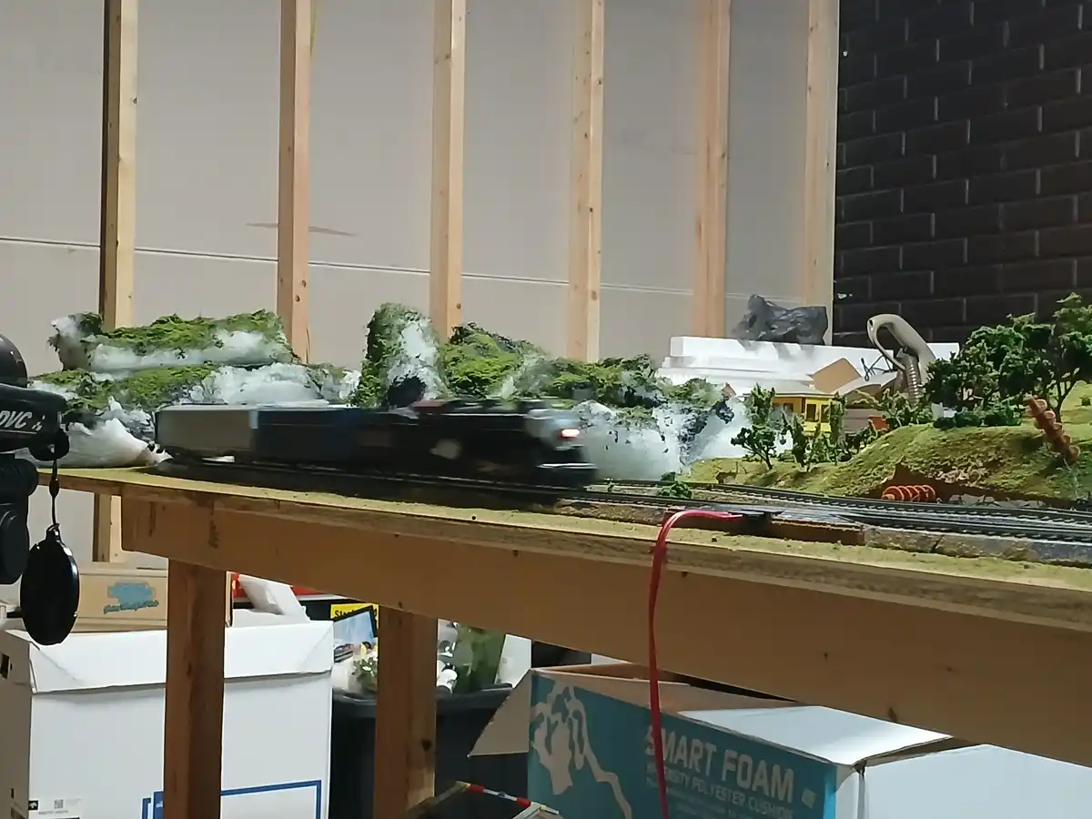 A blurry black model train speeds across a detailed miniature landscape with green hills, white snow patches, and tiny trees…