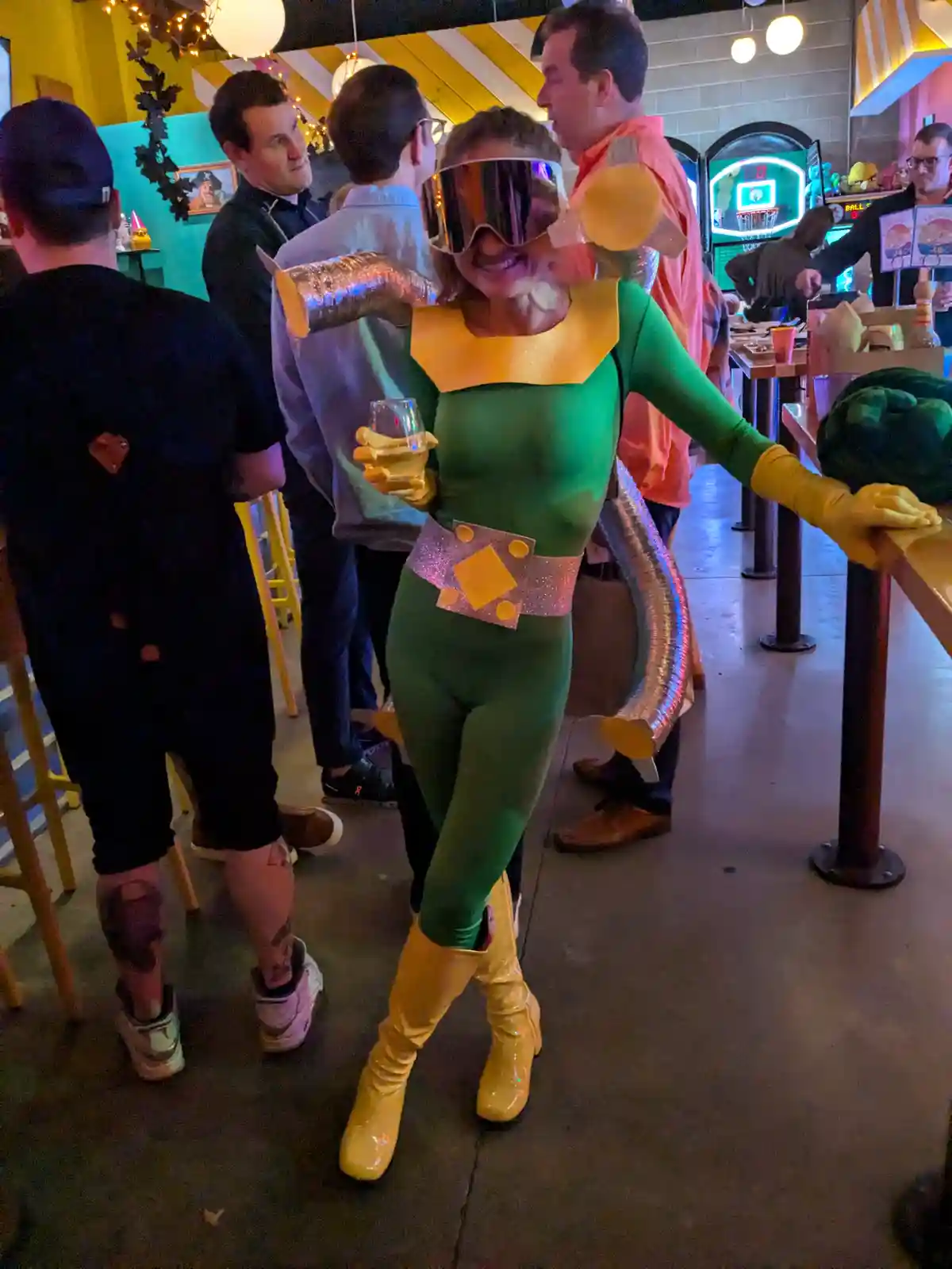 A person in a homemade costume resembling the superhero 'The Tick' stands in a bar, wearing a green bodysuit, yellow boots…