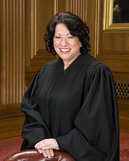 A formal portrait of Justice Sonia Sotomayor, smiling broadly while seated in a courtroom setting. She wears a traditional…