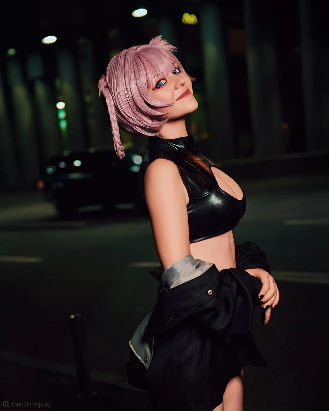 A person with pink hair and blue eyes, wearing a black leather crop top and a black jacket, poses on a city street at night…