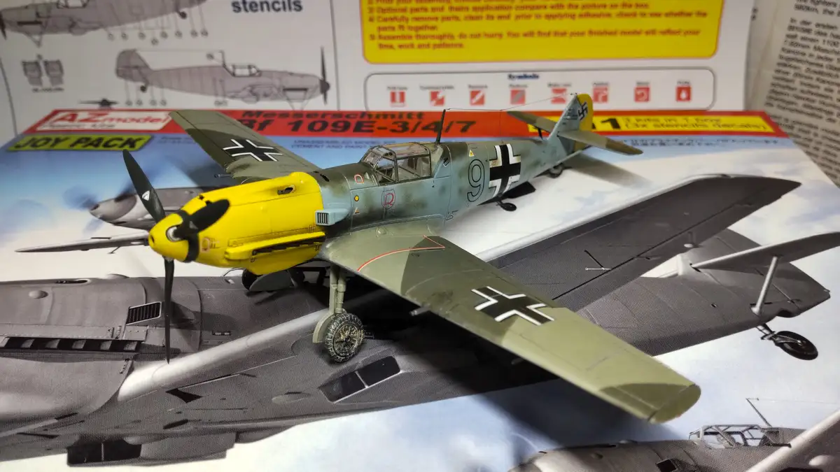 A completed model of a Messerschmitt Bf 109E-3/4/7 fighter plane with a bright yellow nose cone sits atop its instruction…