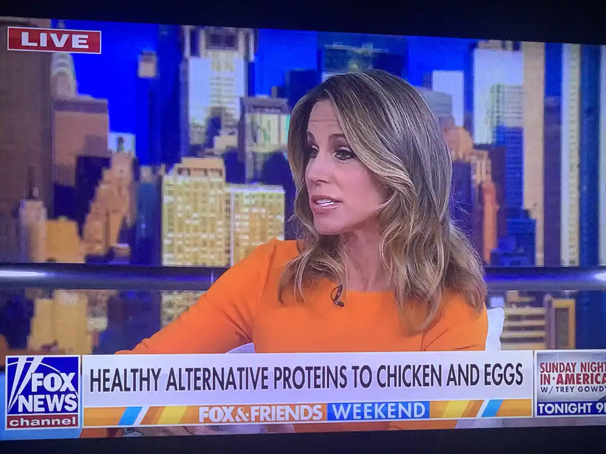 A woman in an orange top is on a Fox News broadcast, with a city skyline backdrop and a 'LIVE' graphic in the top left. The…
