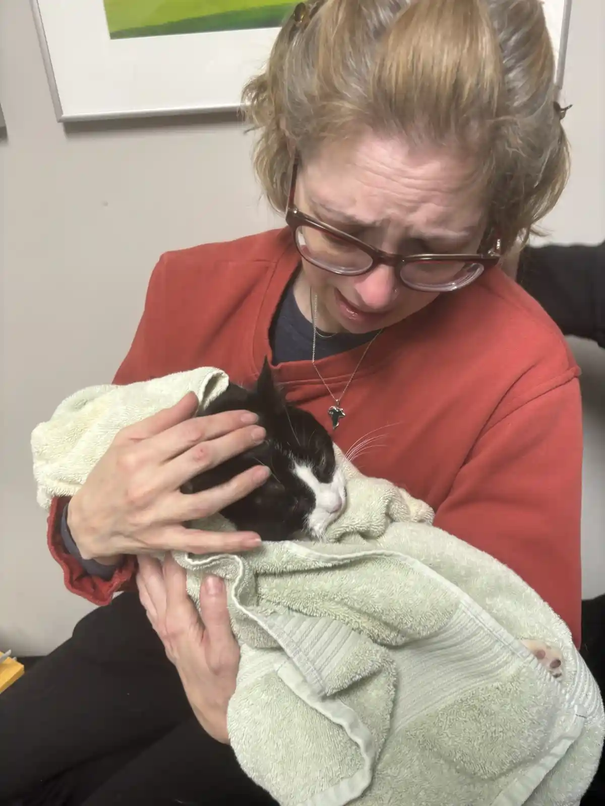 A woman with glasses and reddish-brown hair, wearing a red sweatshirt, is visibly crying while holding a black-and-white cat…