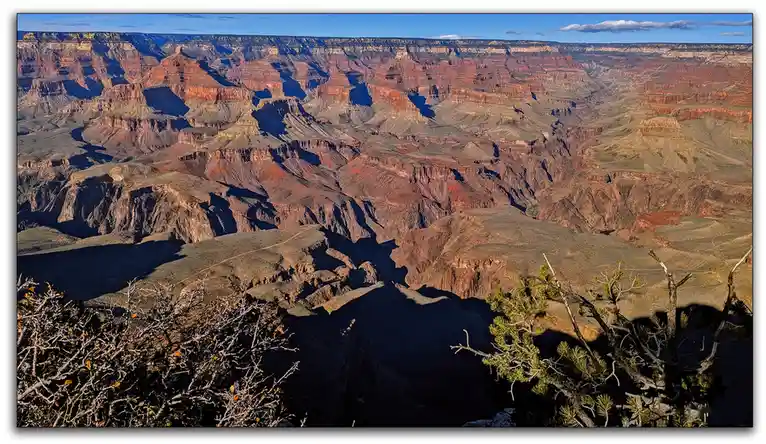 A wide-angle photograph of the Grand Canyon under a clear blue sky, showing layered red rock formations and deep shadows…