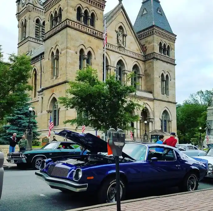A blue classic muscle car with its hood open is parked in front of a large stone courthouse with arched windows and turrets…