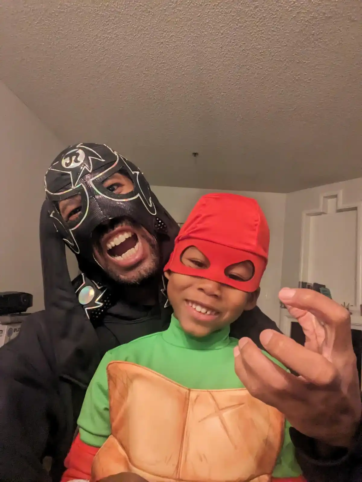 A man in a black lucha libre mask with a wide, toothy grin stands behind a child wearing a red mask and a green shirt with a…