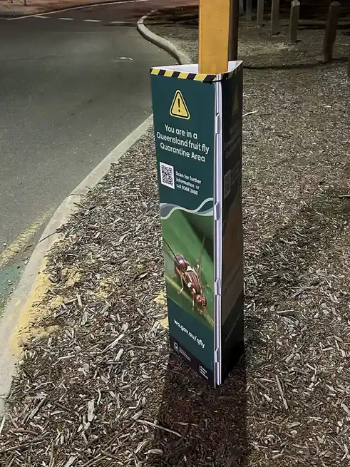 A green public warning sign posted on a pole in a mulched area next to a road, featuring a yellow warning triangle, an image…