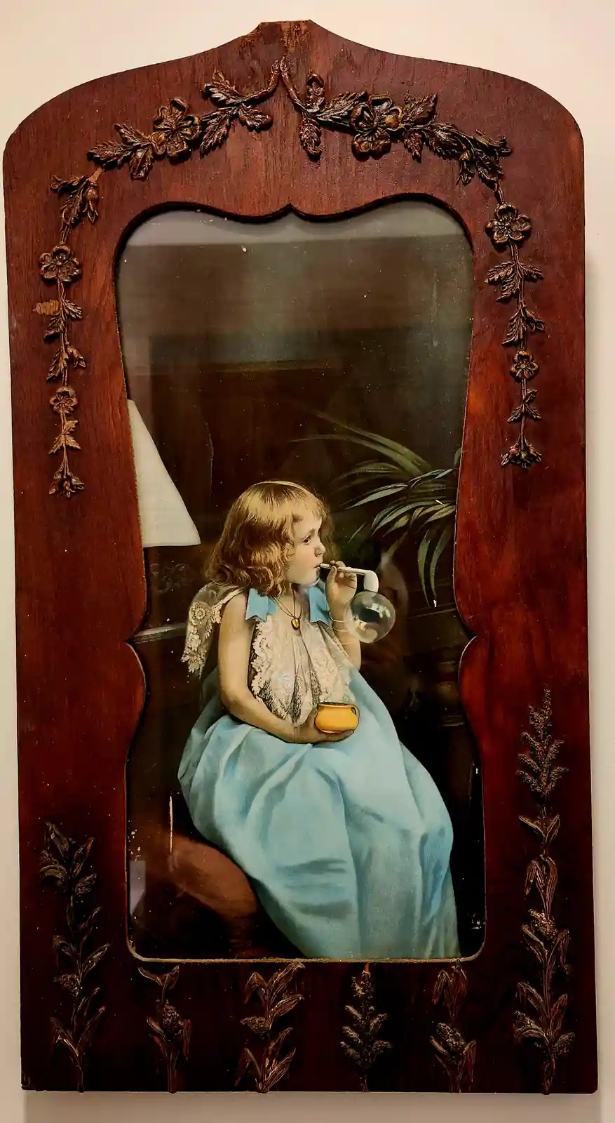 An antique framed painting depicts a young girl in a blue dress blowing bubbles while holding a small yellow cup. The ornate…