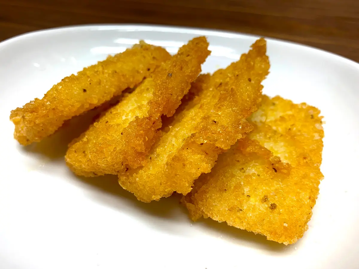 A close-up photo shows several golden-brown, crispy-looking homemade chicken nuggets piled on a white plate, set against a…