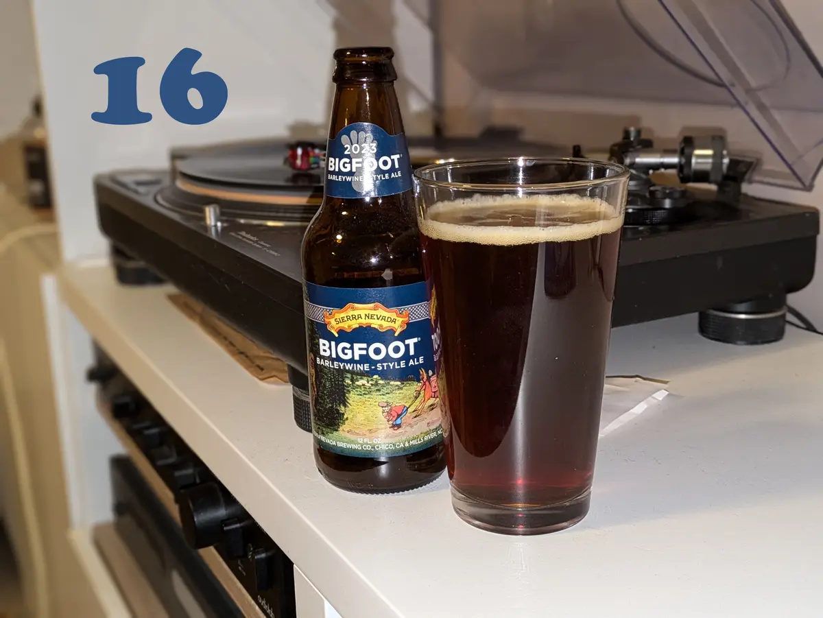 A bottle and glass of Sierra Nevada Bigfoot Barleywine-Style Ale sit on a white countertop in front of a record player. The…