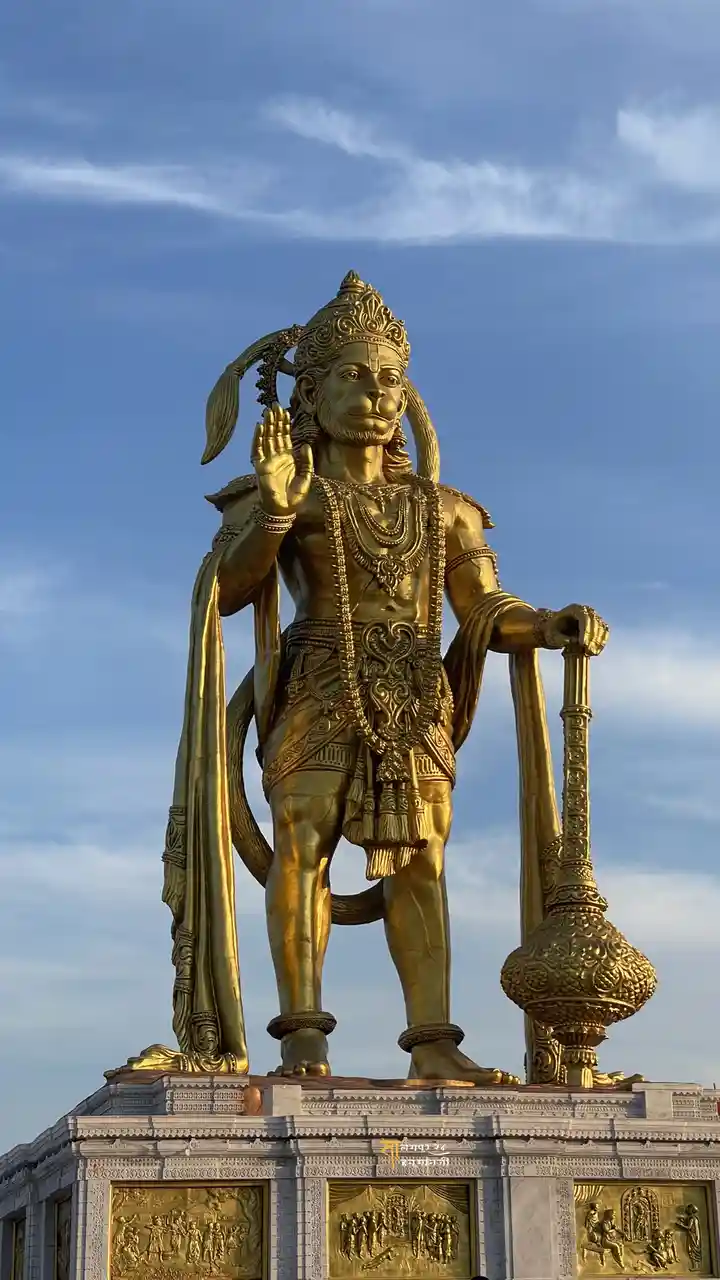 A large golden statue of Hanuman, the Hindu deity with a monkey face, stands against a blue sky with wispy clouds. He wears…