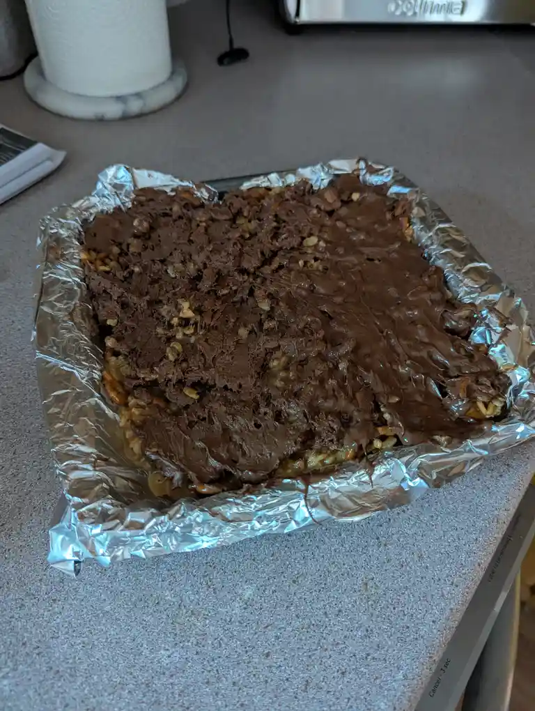 A rectangular baking pan lined with aluminum foil holds a thick, uneven layer of chocolate frosting over a base of what…