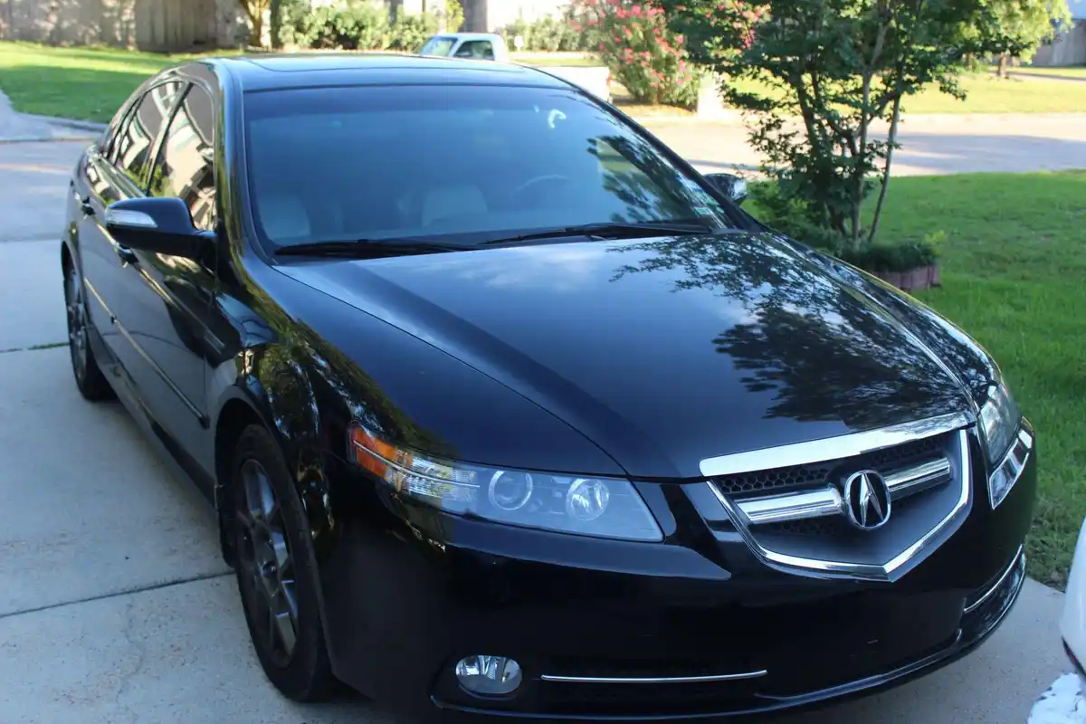 A glossy black Acura TL sedan is parked on a concrete driveway with green grass and trees in the background. The car's front…