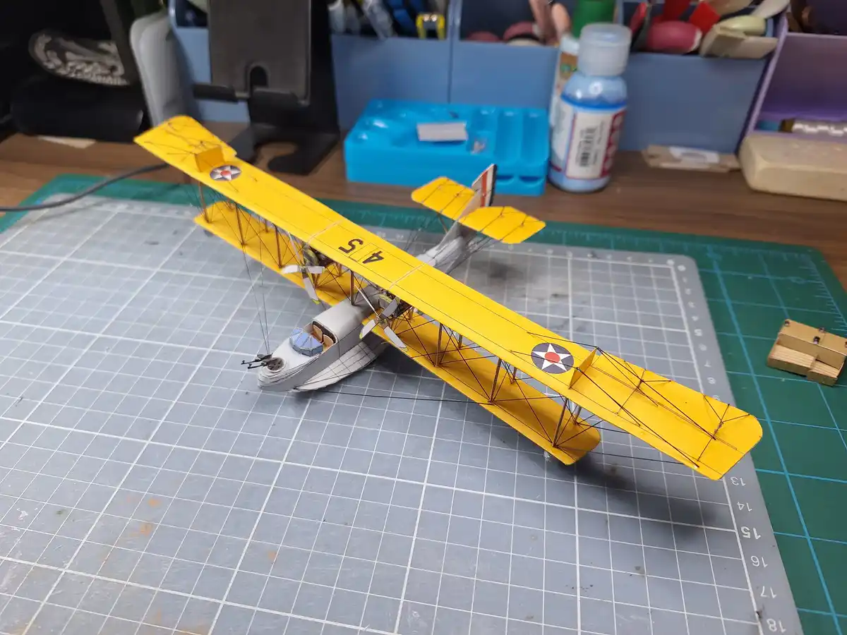 A detailed model of a yellow biplane with a boat fuselage sits on a gray grid cutting mat, surrounded by hobby tools and…
