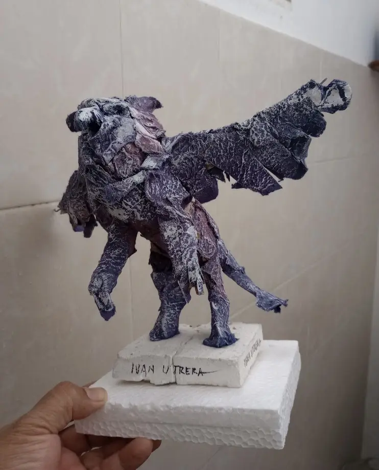 A hand holds a small, textured sculpture of a winged, lion-like creature made from layered paper or fabric, standing on a…