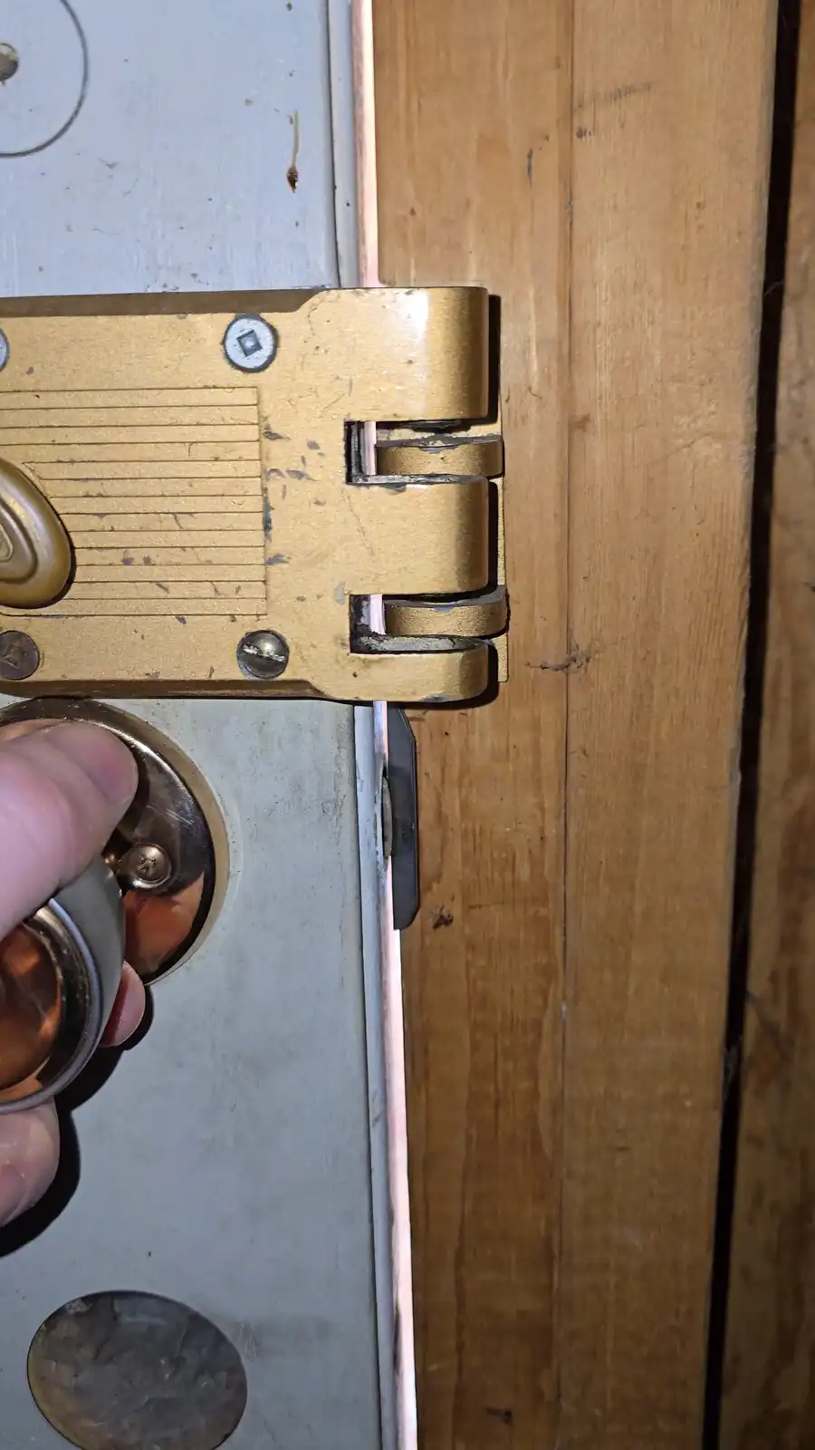 A close-up photo shows a person's thumb pressing a door handle on a light gray metal door, with a brass-colored latch…