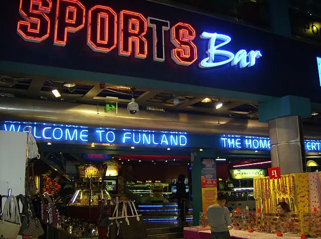 A photo of a brightly lit storefront with a large neon sign reading 'SPORTS Bar' in red and blue. Below it, a blue neon sign…