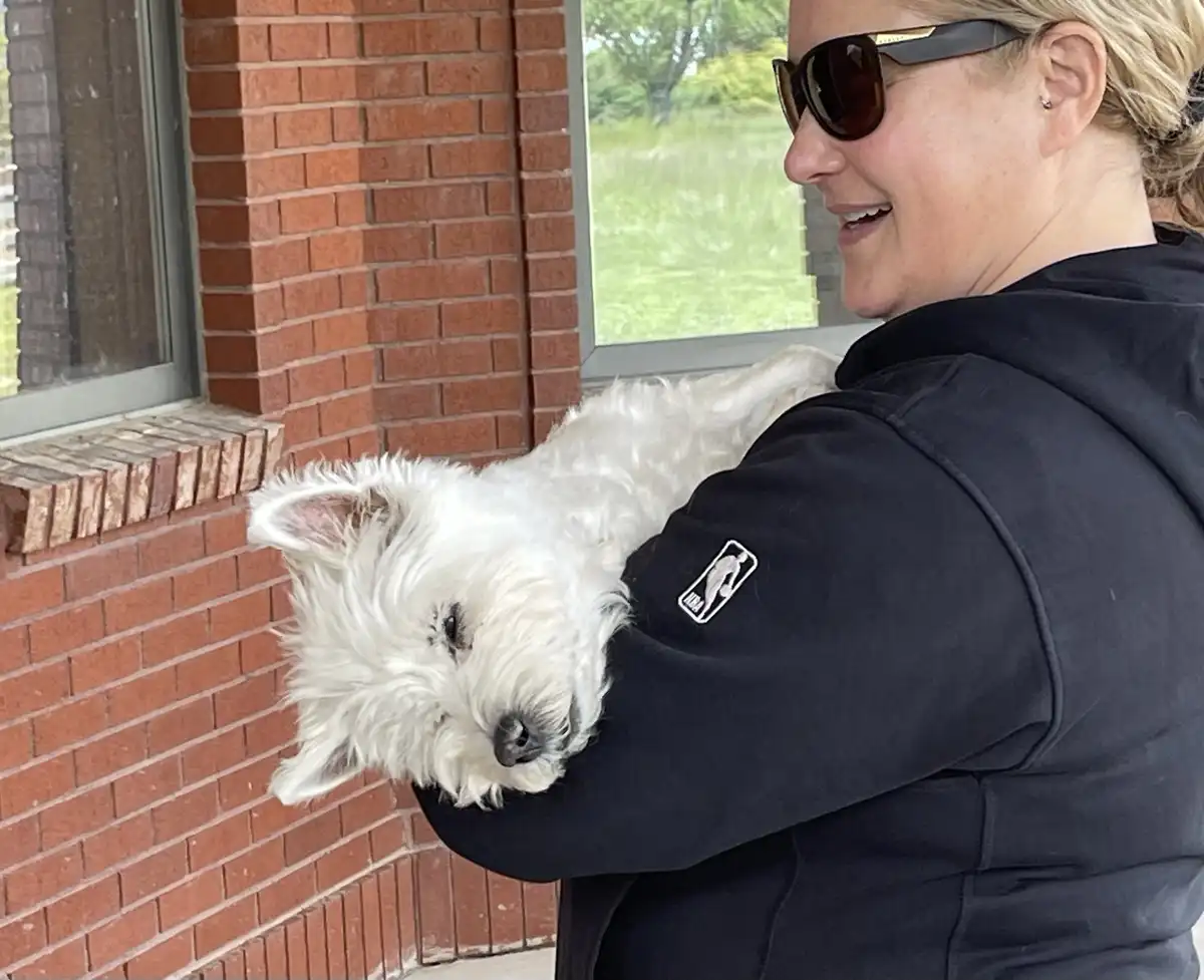 A woman wearing sunglasses and a black hoodie holds a small white dog, which is resting its head on her arm with its eyes…