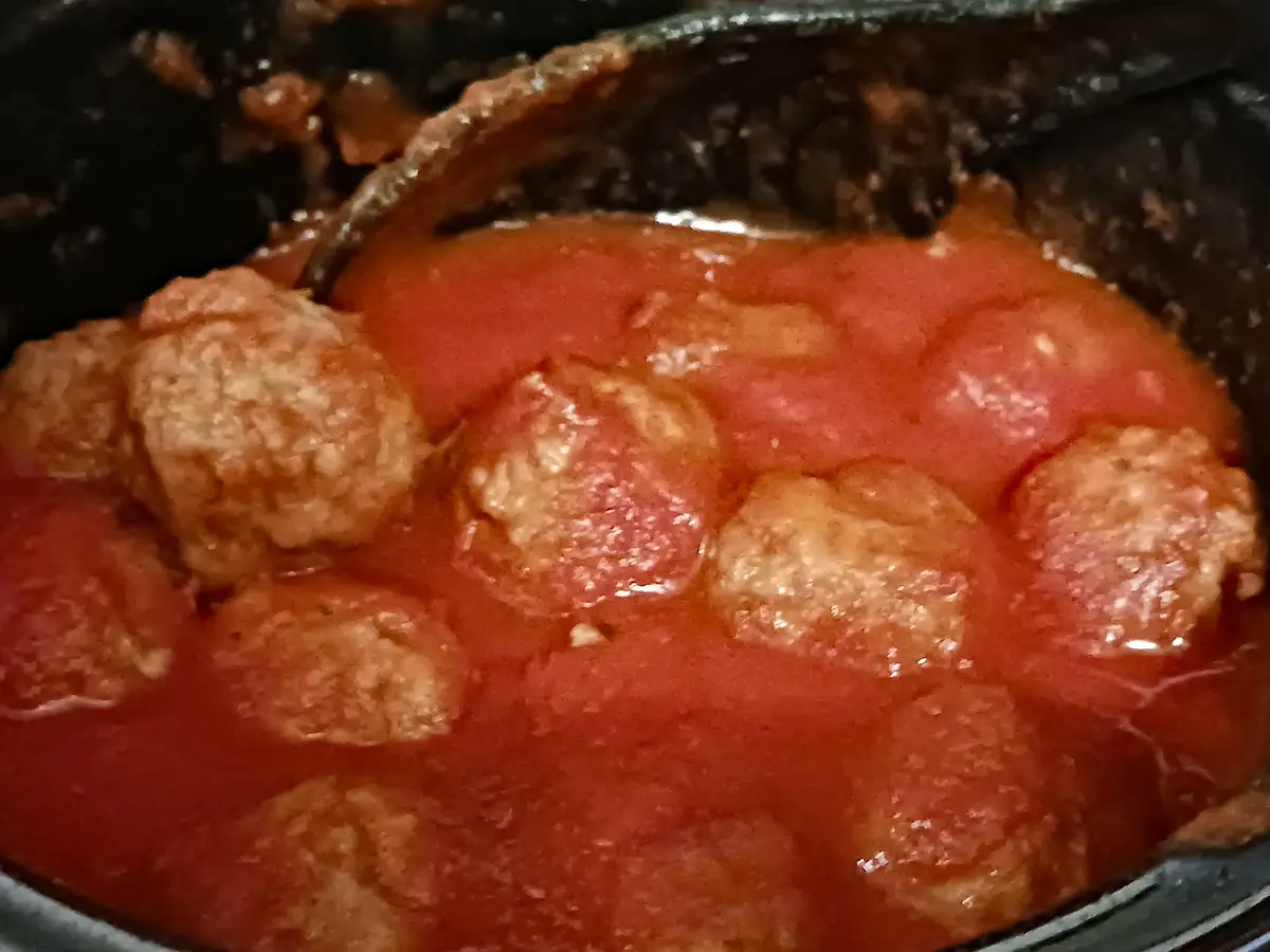A close-up photo shows several brown meatballs partially submerged in a thick, red tomato sauce inside a dark cooking pot…
