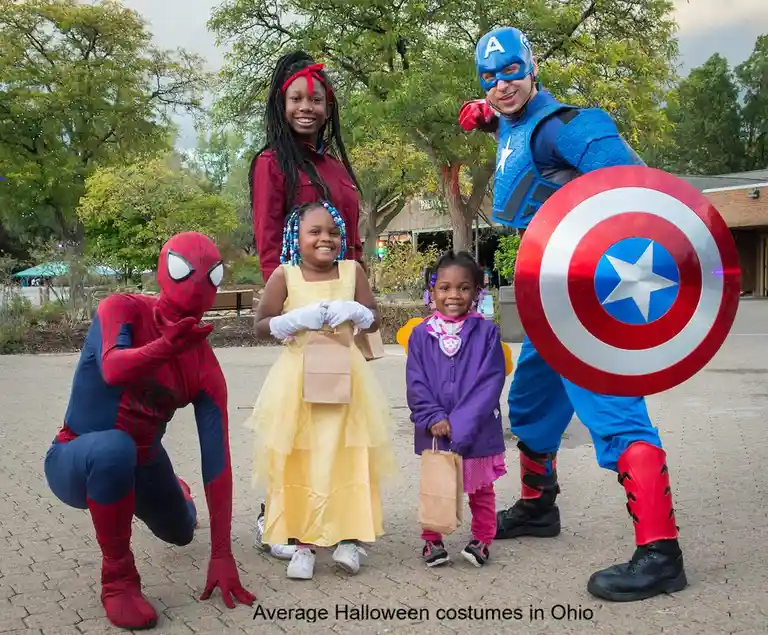 A family of four poses outdoors in costumes: a man as Spider-Man crouching, a woman in a red jacket, a young girl in a…