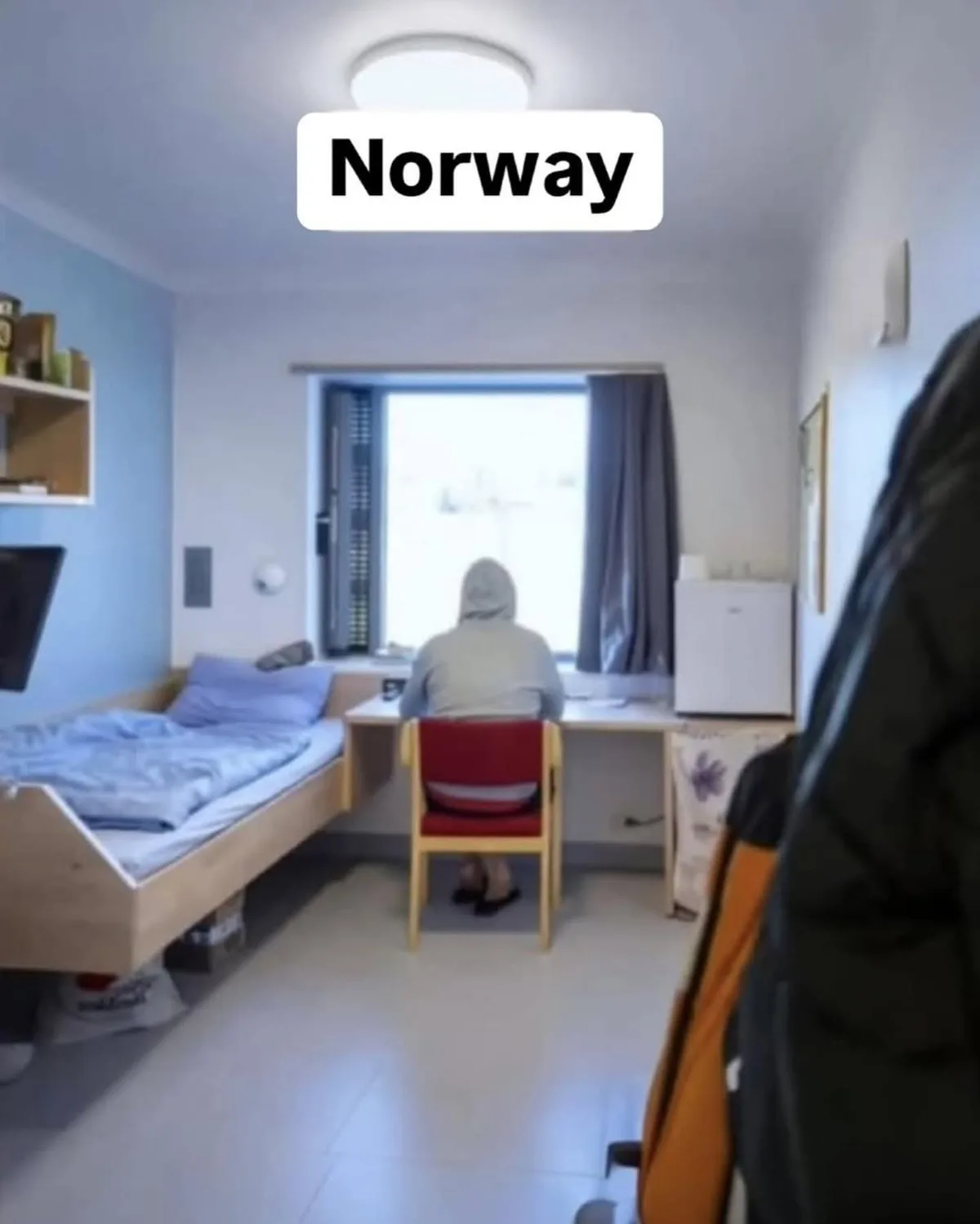 A person in a grey hoodie sits at a desk in a minimalist, brightly lit room with a single bed, facing a large window. The…