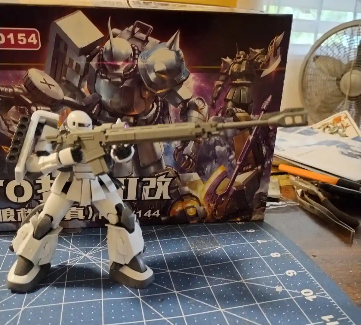 A white plastic model of a Gundam-style robot stands on a blue grid cutting mat, holding a long rifle. Behind it is its…